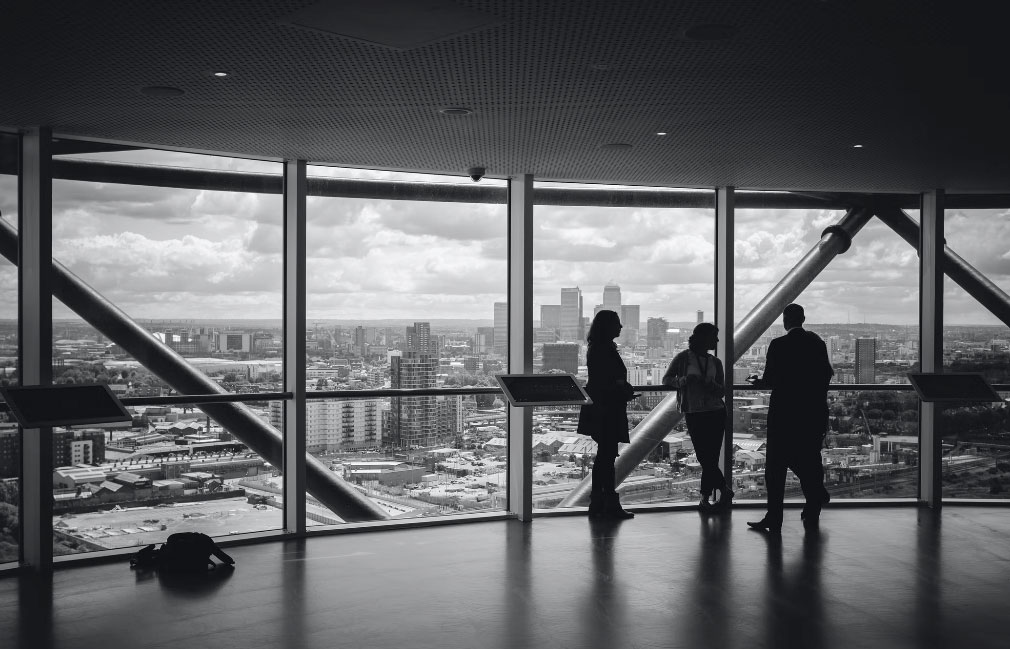 Three people standing near large windows talking