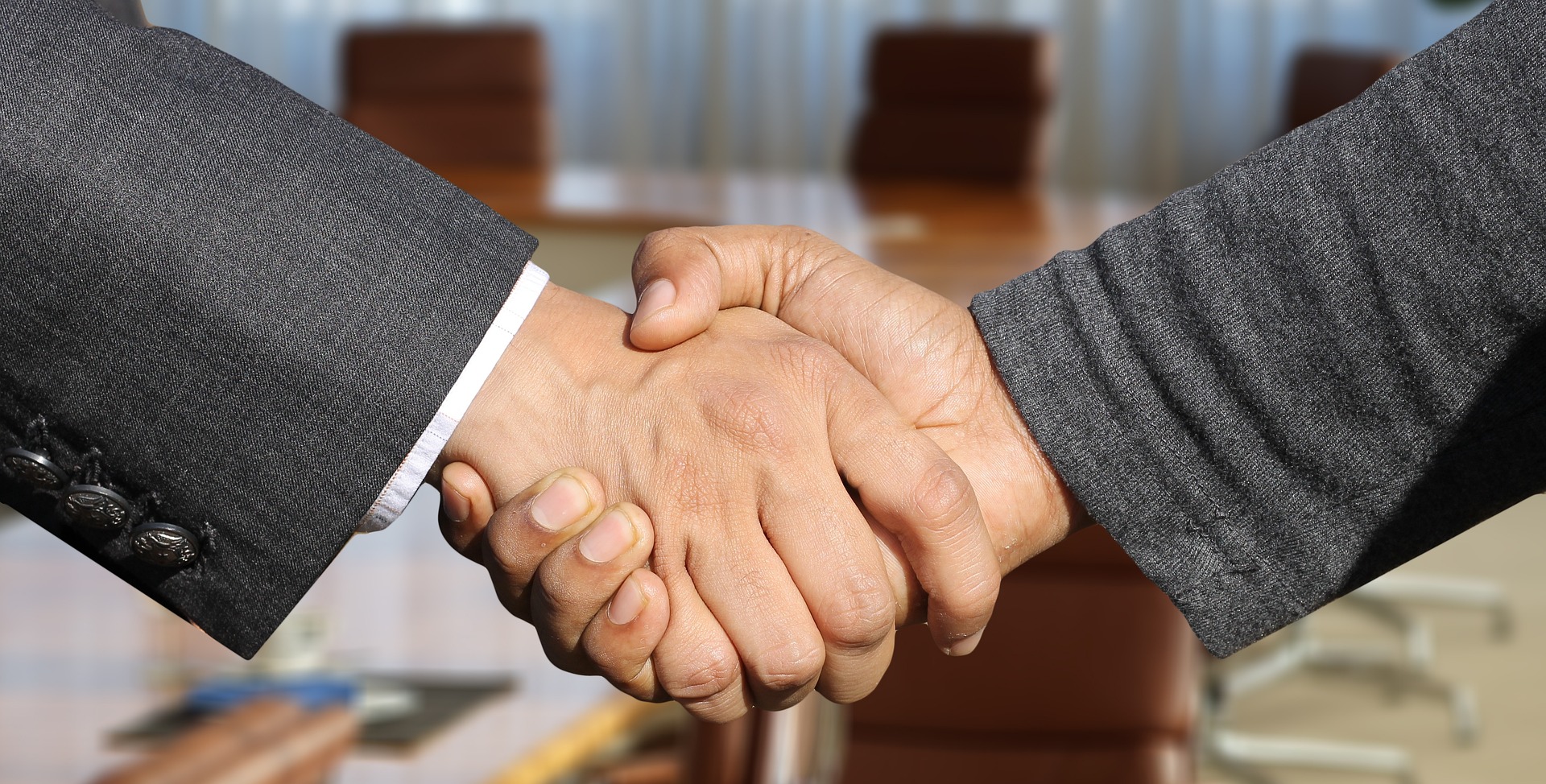 An image of a handshake by two men in grey suits. Just their arms and hands are visible.