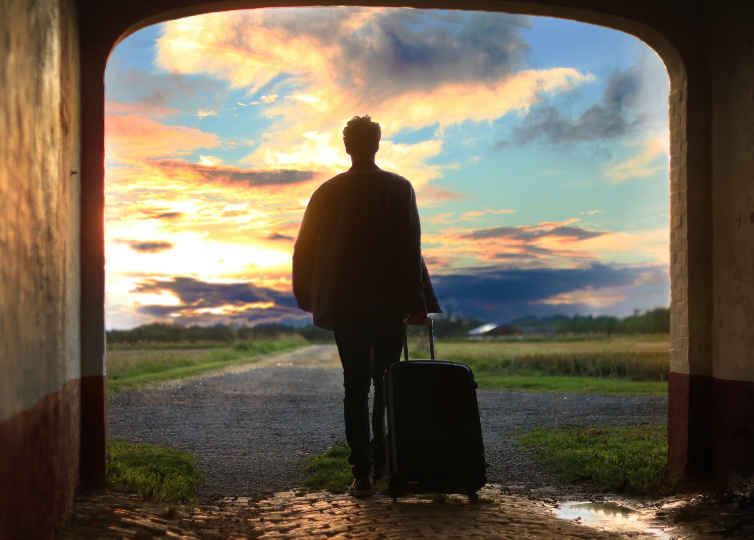 man with a carry-on case standing in an archway looking out onto a sunset