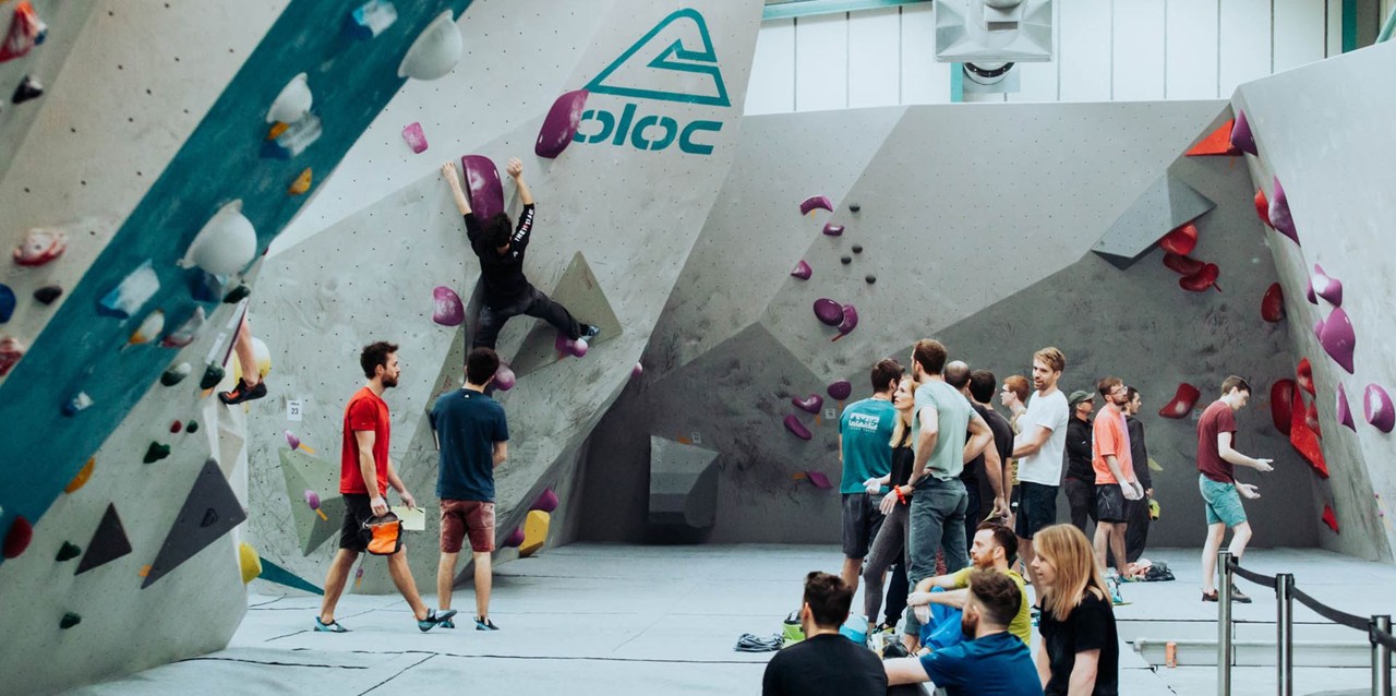 A white bouldering wall with climbers hanging from climbing holds and a group standing watching